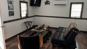 Un salon avec un canapé en cuir noir, une table basse en bois, une petite table d’appoint et un téléviseur à écran plat fixé au mur.