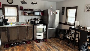 Una cocina con armarios de madera, electrodomésticos de acero inoxidable, un microondas y un refrigerador. Hay taburetes de bar y una pequeña mesa de comedor.