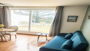 Una sala de estar con un sofá azul, una mesa de comedor con sillas, una pequeña mesa redonda y un amplio ventanal corredizo que da a una terraza exterior.