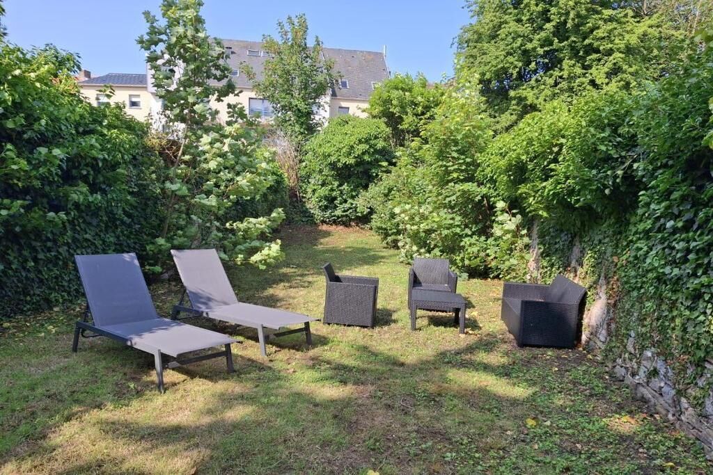 A garden with outdoor furniture, including lounge chairs and a table, surrounded by greenery and a building in the background.. Spa Sauna et 35 m2 charmant plus 30 m2 de terrasse