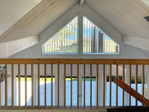 A room with a sloped ceiling, wooden beams, a balcony with a railing, and a view of a body of water and greenery outside. - Bright house with sea view at Lindö (Listerby)