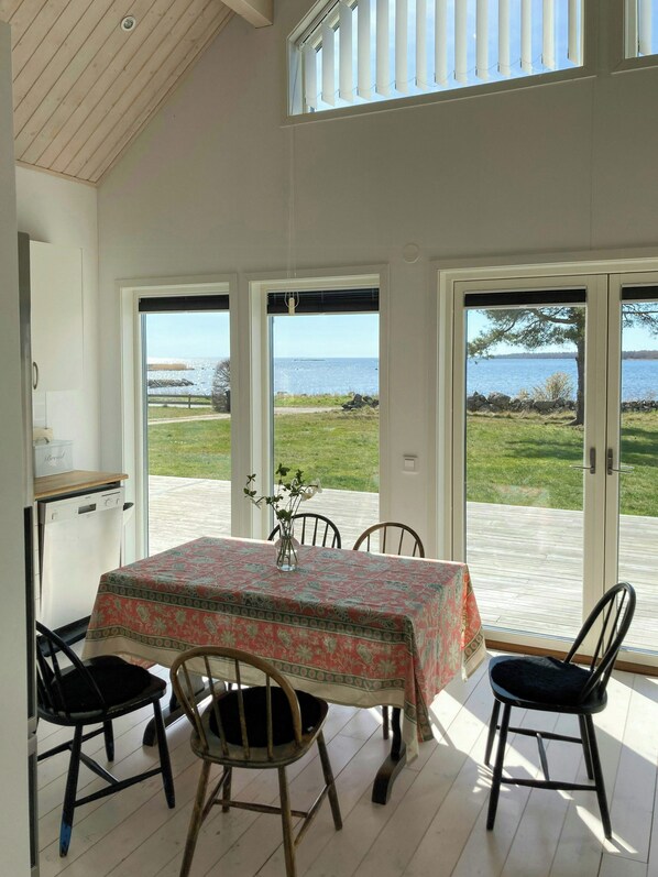 Un coin repas avec une table dressée pour quatre personnes, de grandes fenêtres offrant une vue sur la mer et un plafond orné de poutres en bois.