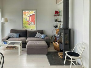 A living room with a sofa, a table with a laptop, a bookshelf, a fireplace, and a view of a red house outside. - Bright house with sea view at Lindö (Listerby)