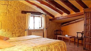 A bedroom with a stone wall, wooden beams, a bed, a window with curtains, a table, and a chair. - La Casa de las Monjas (Miranda de Arga)