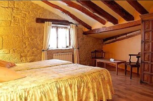 Un dormitorio con pared de piedra, vigas de madera, cama, ventana con cortinas, una mesa y una silla.