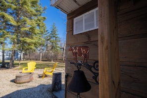 A wooden cabin with a bell and a cow decoration on the wall, a chair, and a fire pit in the yard.
