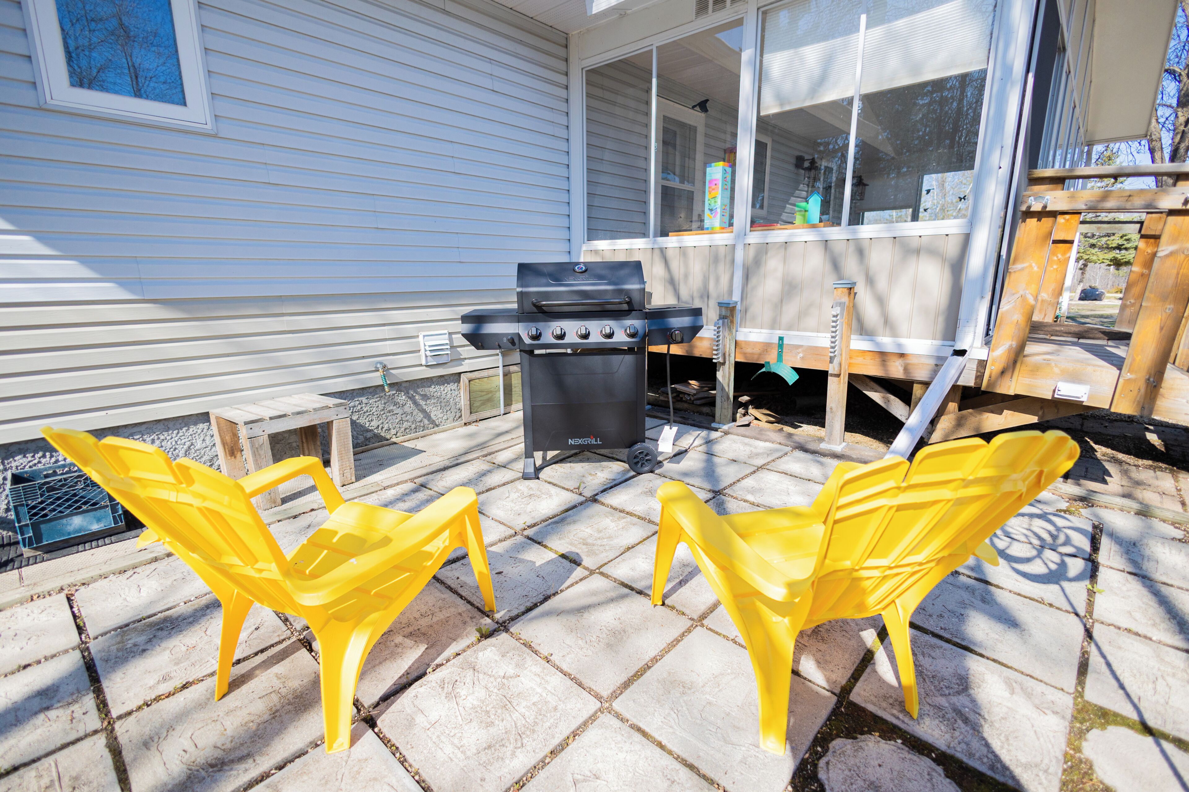 Een terras met een barbecue, twee gele stoelen en een houten tafel.