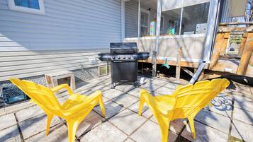 Een terras met een barbecue, twee gele stoelen en een houten tafel.