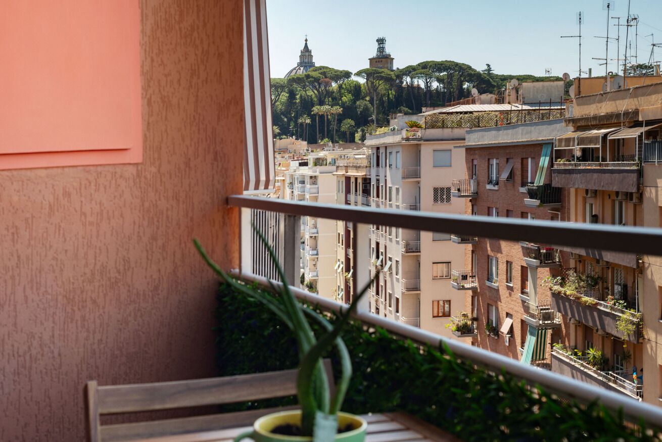 Un balcone con una pianta in vaso, vista su un paesaggio urbano con edifici e alberi, e cielo sereno.