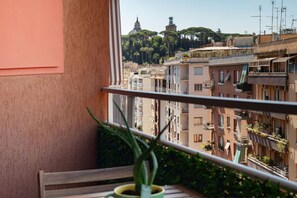Un balcón con una planta en maceta, vista de un paisaje urbano con edificios y árboles, y cielo despejado.