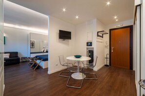 Ein modernes Zimmer mit einem runden Esstisch, transparenten Stühlen, einem Flachbildfernseher, einer Holztür und einer kleinen Küchenzeile.