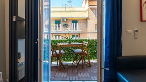 Een open deur die leidt naar een balkon met een tafel, stoelen, een plant in pot en uitzicht op een gebouw met balkons.
