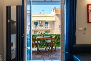 Eine geöffnete Tür führt auf einen Balkon mit Tisch und Stühlen, einem Blumentopf und Blick auf ein Gebäude mit Balkonen.