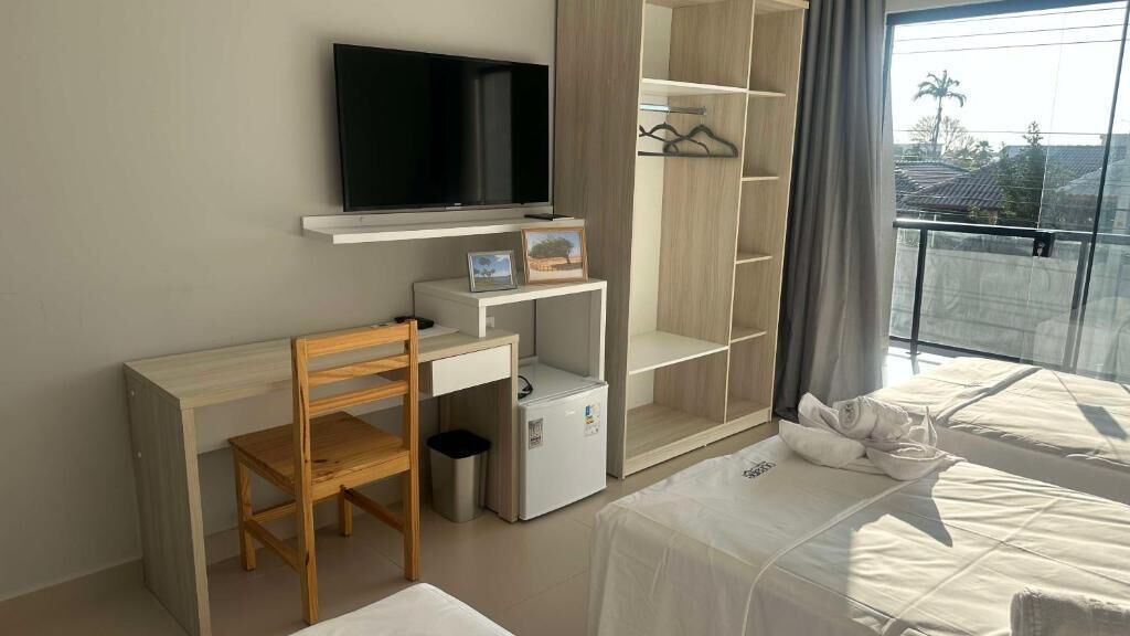 Un dormitorio con cama, escritorio, silla, televisor, armario y balcón con vistas.