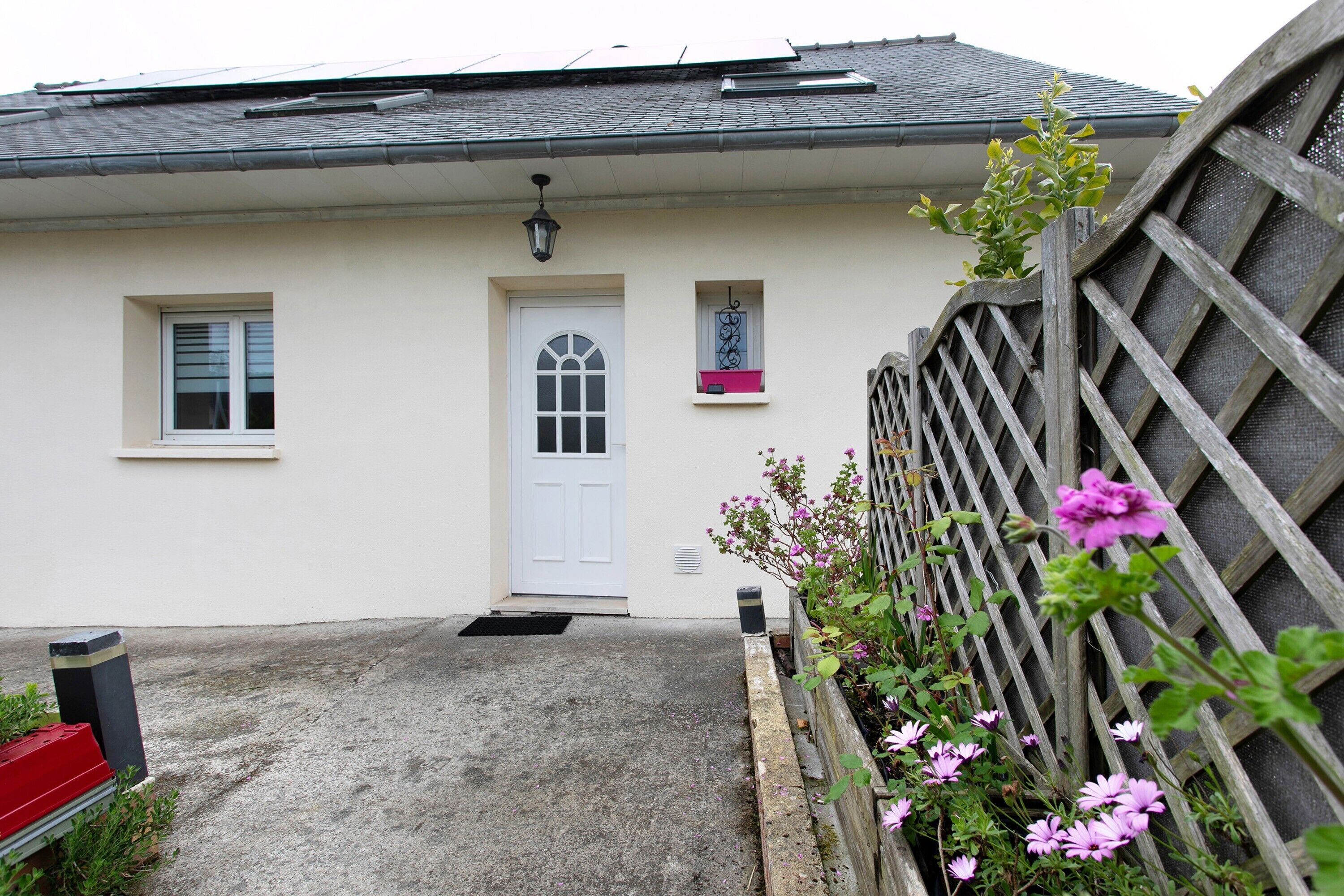 A house with a white exterior, a front door, two windows, a small balcony, and a wooden fence with flowers.