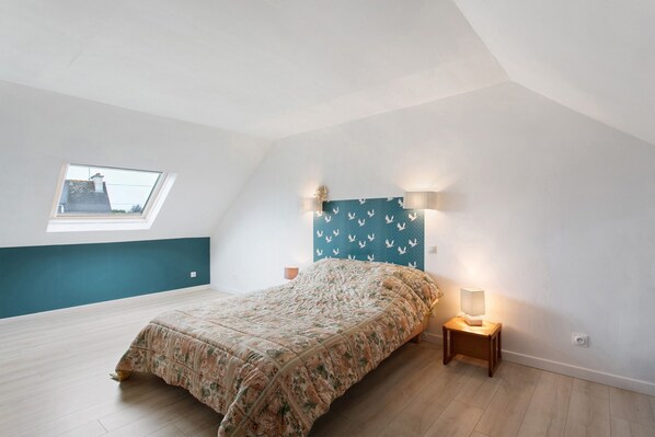 Ein Schlafzimmer mit schrägem Dach, einem Oberlicht, einem Bett mit floralem Überwurf, zwei Wandleuchten und einem kleinen Nachttisch.