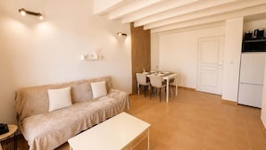 A modern living room with a sofa, a dining table with chairs, a small kitchen area with a refrigerator, and a door leading to another room. - La parenthèse Camarguaise (Gallargues-le-Montueux)