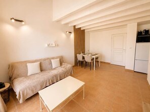 Una sala moderna con un sofá, una mesa de comedor con sillas, una pequeña cocina con un refrigerador y una puerta que da a otra habitación.