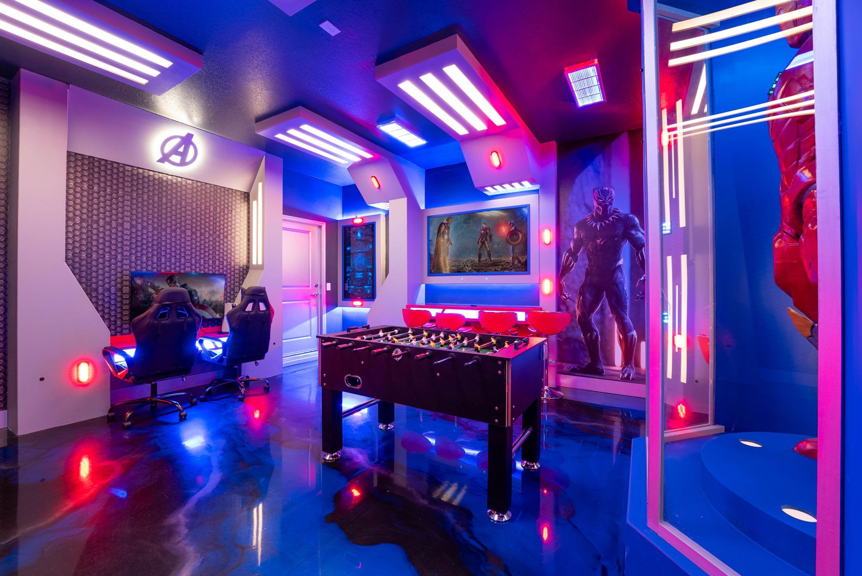 Una habitación con mesa de fútbol de mesa, sillas tipo gamer, una pantalla grande y una pared con un logotipo.