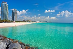 Ein Strand mit klarem türkisblauem Wasser, einem sandigen Ufer und Hochhäusern im Hintergrund.
