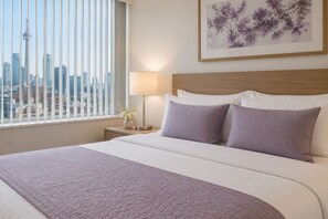 Ein Hotelzimmer mit Bett, Nachttisch, Lampe, einem gerahmten Blumenbild und einem Fenster mit Blick auf die Stadt.