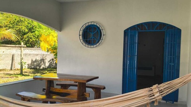 A hammock, a wooden table, and a blue door in a room with a tiled floor and a wall-mounted fan. - Grandma's house in Piúma: Backyard, Porch and 3 Bedrooms (Piúma)
