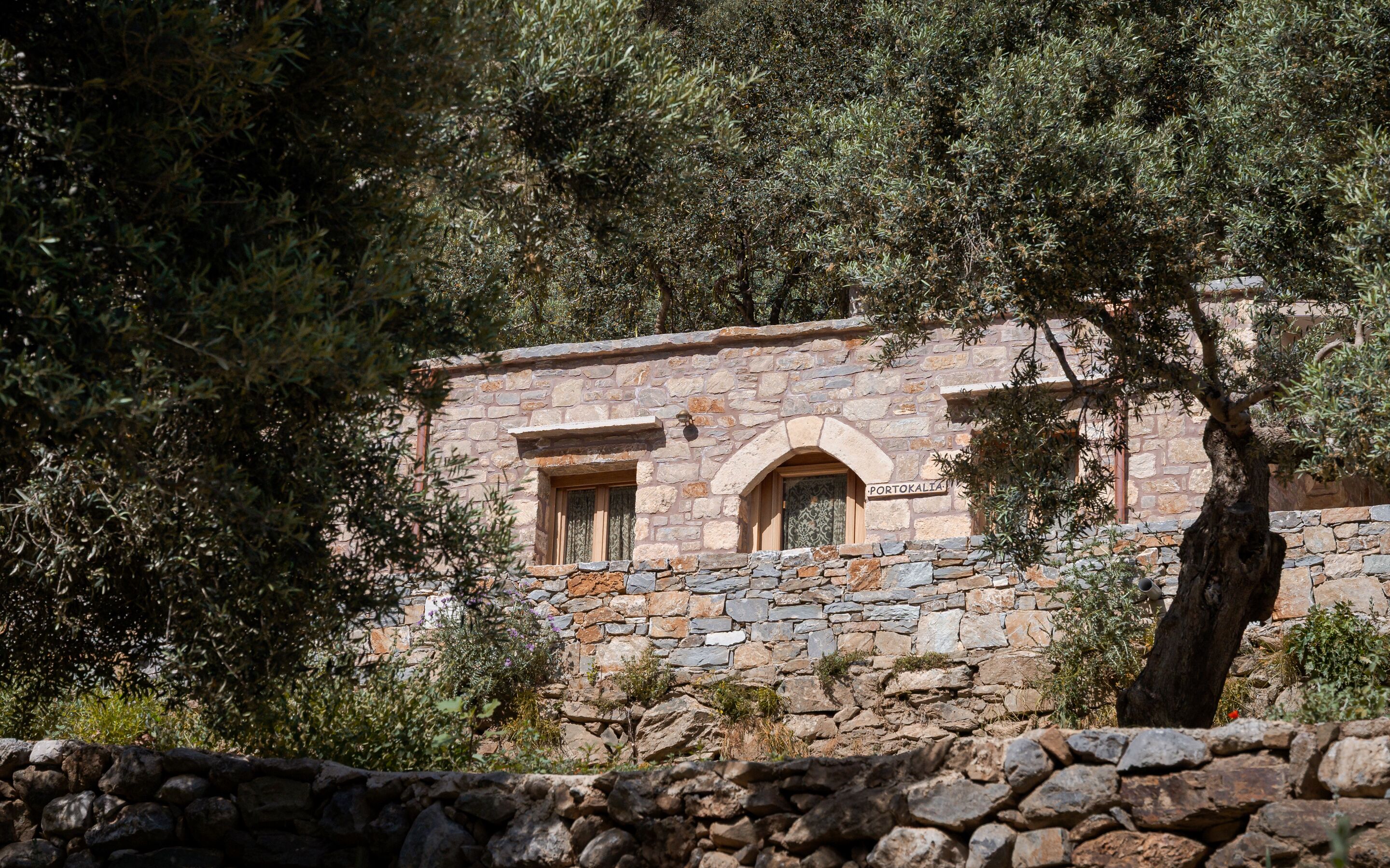 Un edificio de piedra con dos ventanas, un muro de piedra al frente y árboles al fondo.