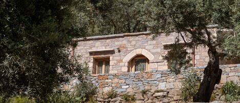 Un edificio de piedra con dos ventanas, un muro de piedra al frente y árboles al fondo.