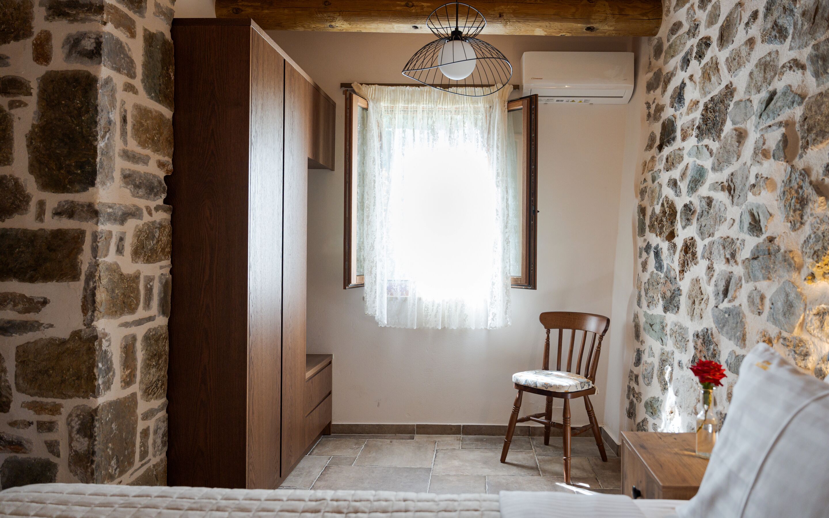 Une pièce aux murs de pierre, avec une chaise en bois, un lit et une fenêtre ornée d’un rideau.