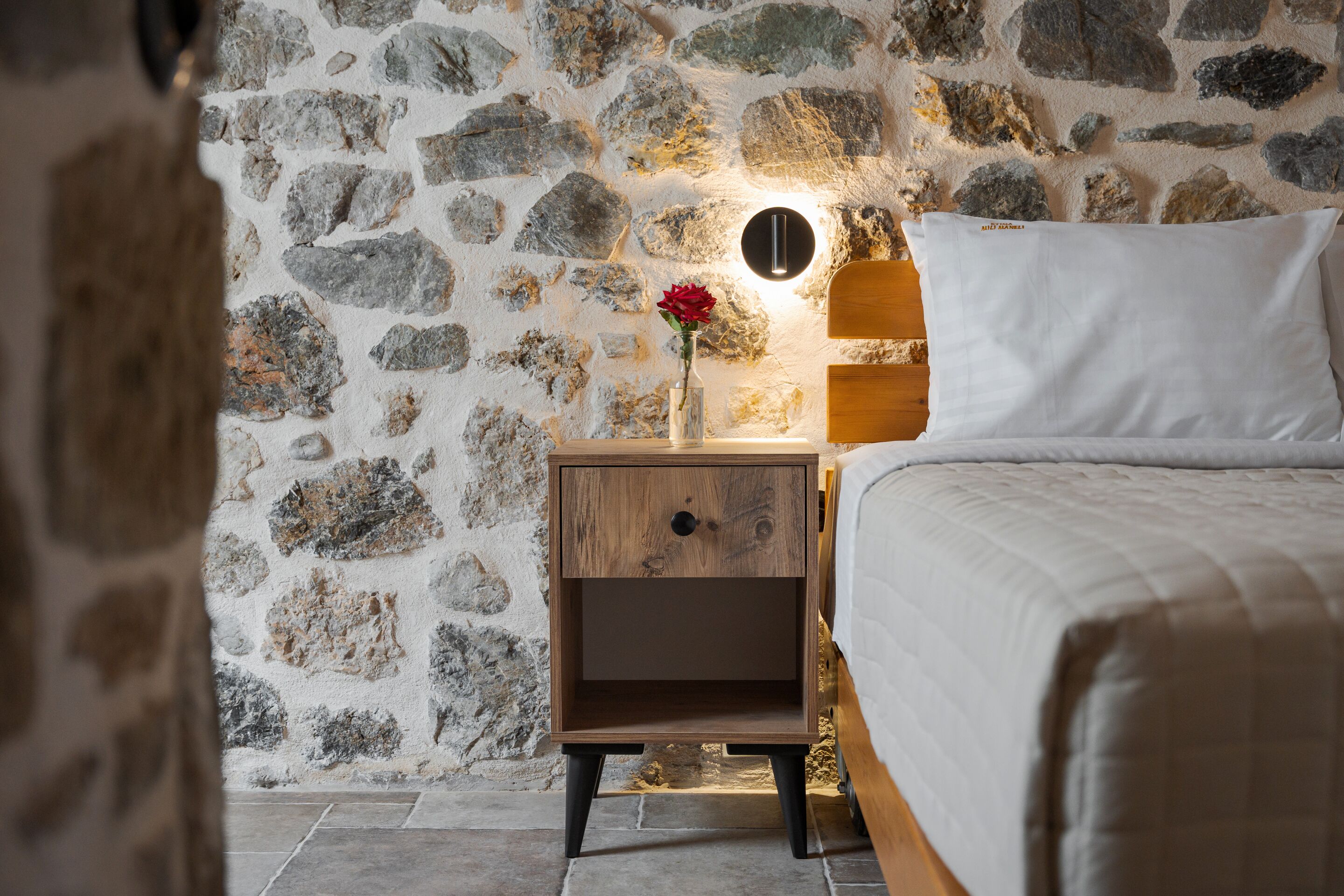 A bedroom with a stone wall, a wooden nightstand with a vase of flowers, and a bed with white bedding.