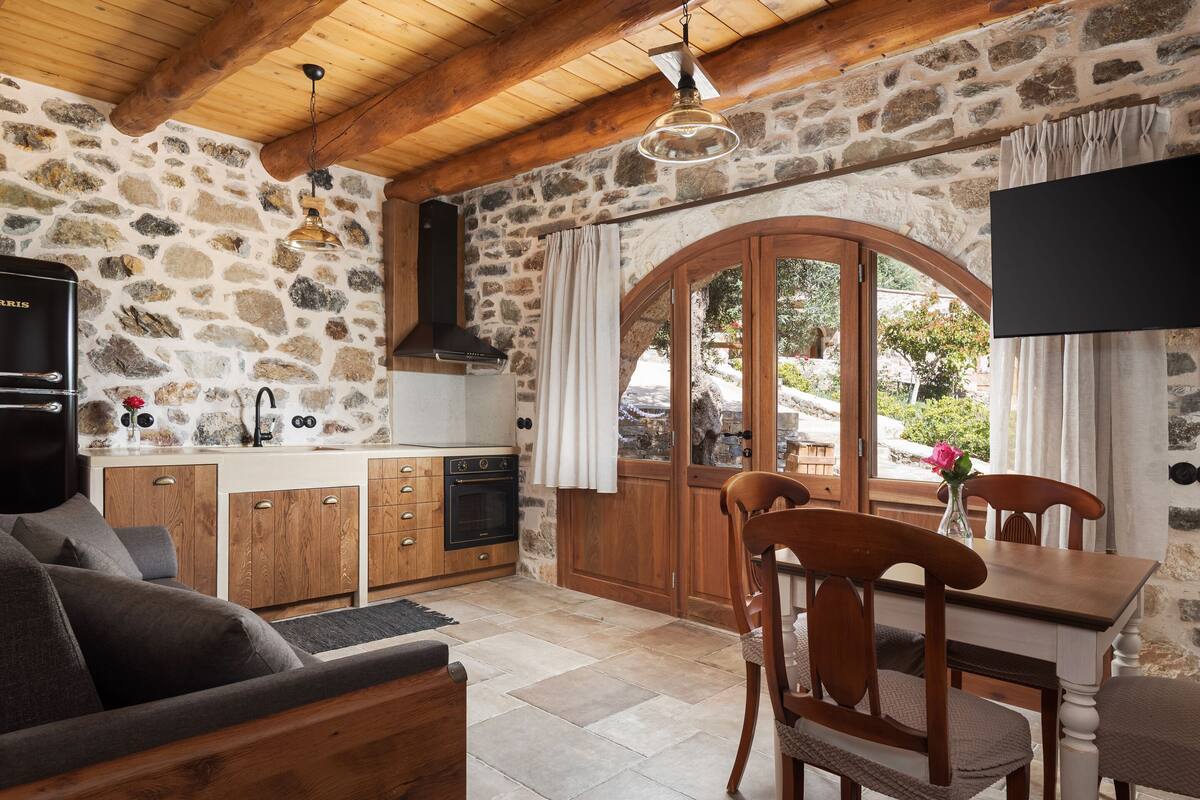 Una cocina rústica con paredes de piedra, vigas de madera en el techo, un horno empotrado y una zona de comedor con mesa y sillas de madera.