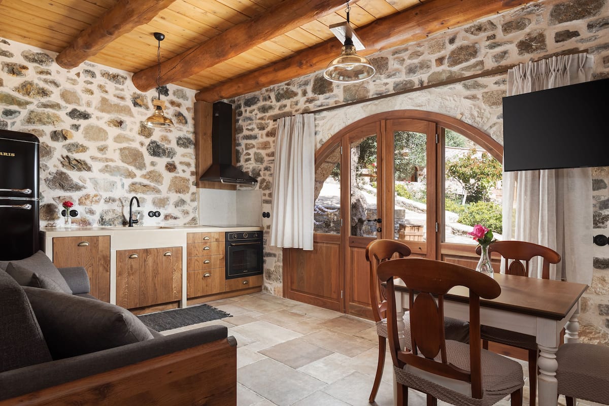 Une cuisine rustique aux murs en pierre, avec des poutres en bois au plafond, un four encastré et un coin repas comprenant une table et des chaises en bois.