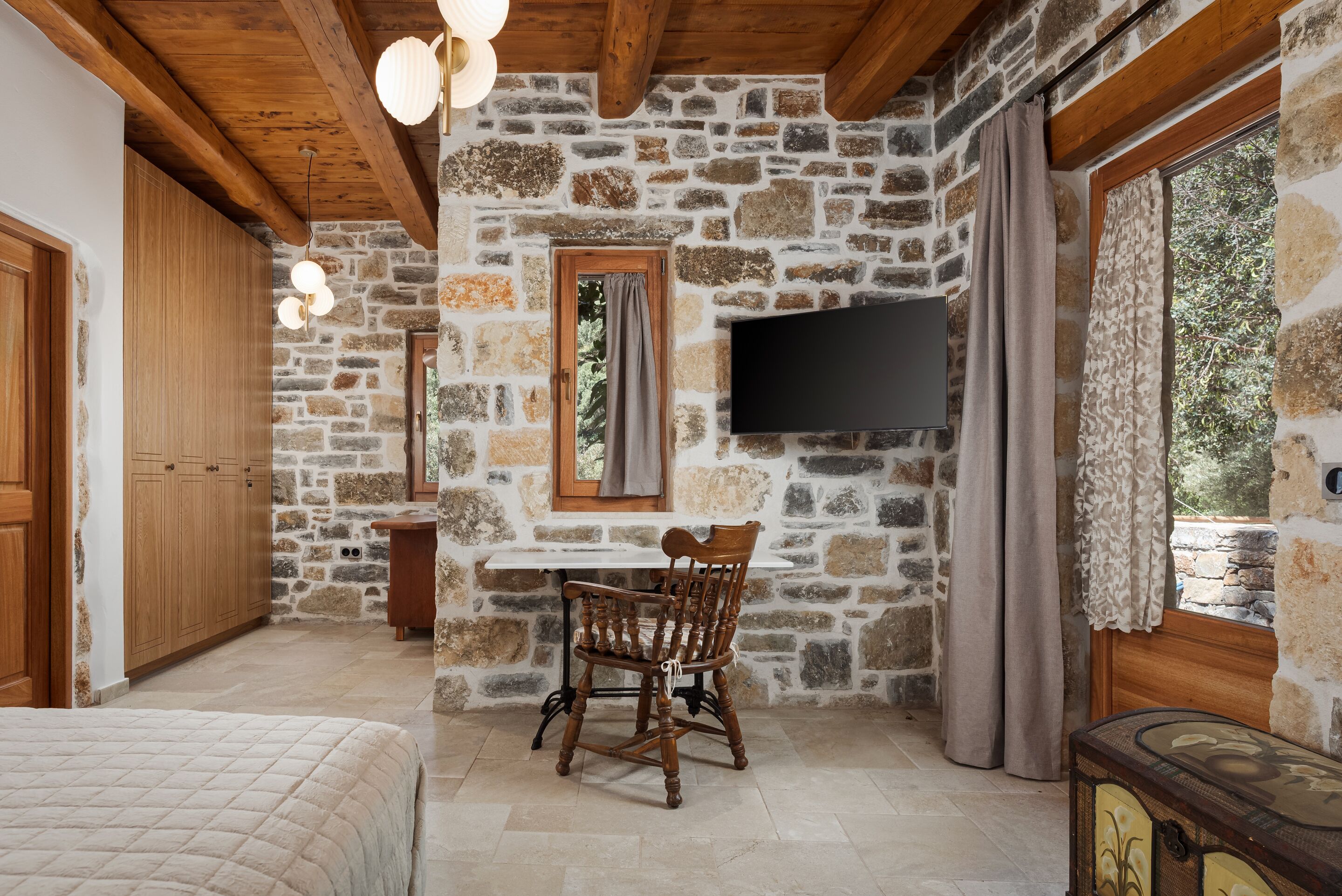 Une pièce aux murs de pierre et au plafond en bois, avec un téléviseur fixé au mur, une chaise en bois et un lit recouvert d’une courtepointe matelassée.