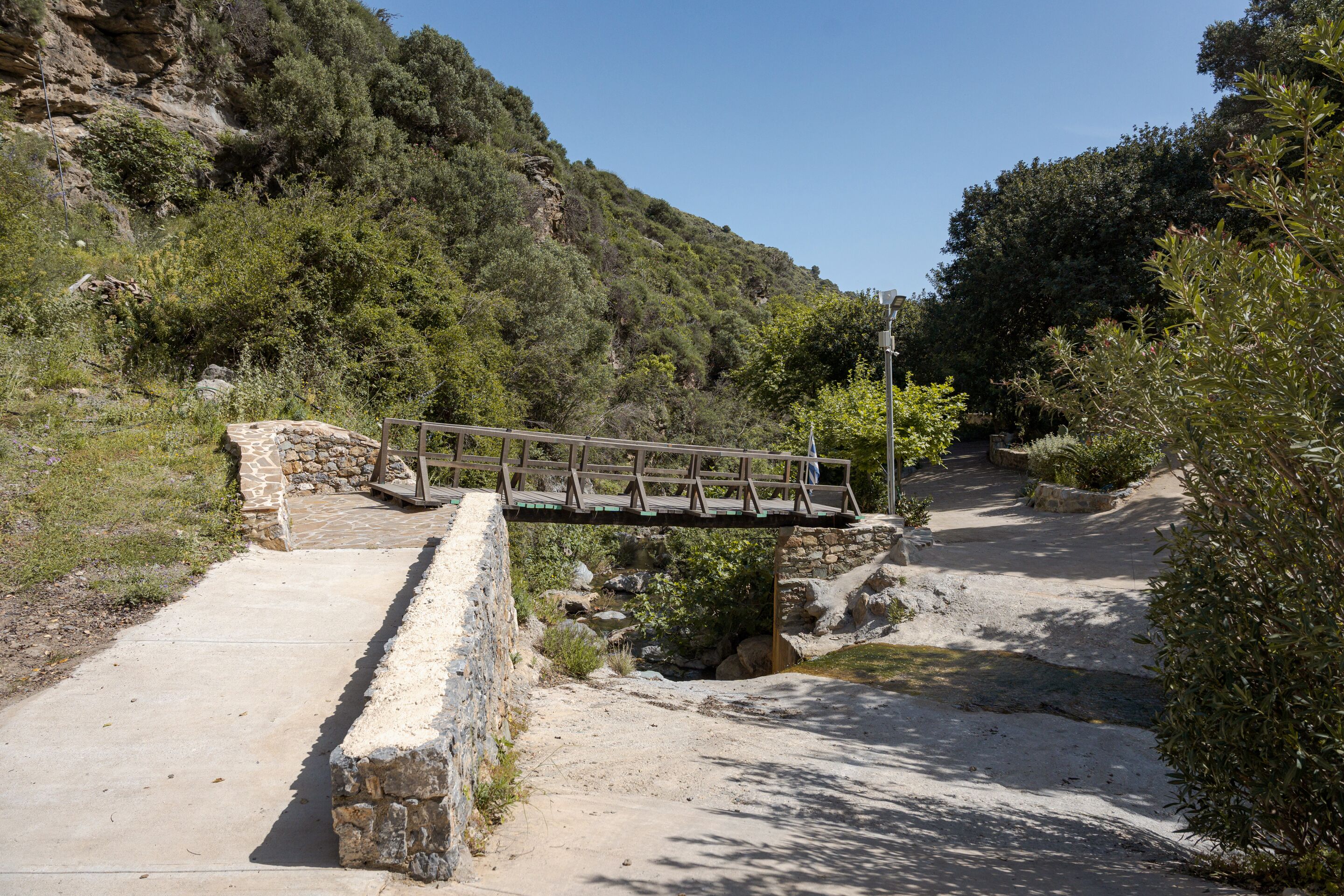 A stone bridge over a concrete path in a natural setting with greenery and a rocky hillside.