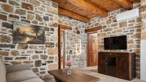 A stone-walled room with a wooden ceiling, a TV on a cabinet, a sofa, and a table with a vase. - Manelis Mills Estate - Guest Houses (Myrthianos Plakias)