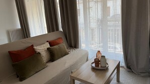 Sala de estar con un sofá, almohadas, una mesa con bandeja y vistas a un edificio a través de cortinas translúcidas.