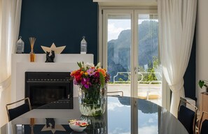 A modern dining room with a glass table, a fireplace, a vase of flowers, and a balcony with a view of mountains.