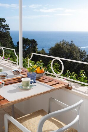 A wooden table with a white chair, a glass of water, a cup, and a bowl of fruit on a balcony overlooking the sea.
