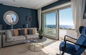 Un salon moderne avec un canapé, une table basse et un fauteuil, une grande fenêtre donnant sur l’océan et un miroir rond accroché au mur.