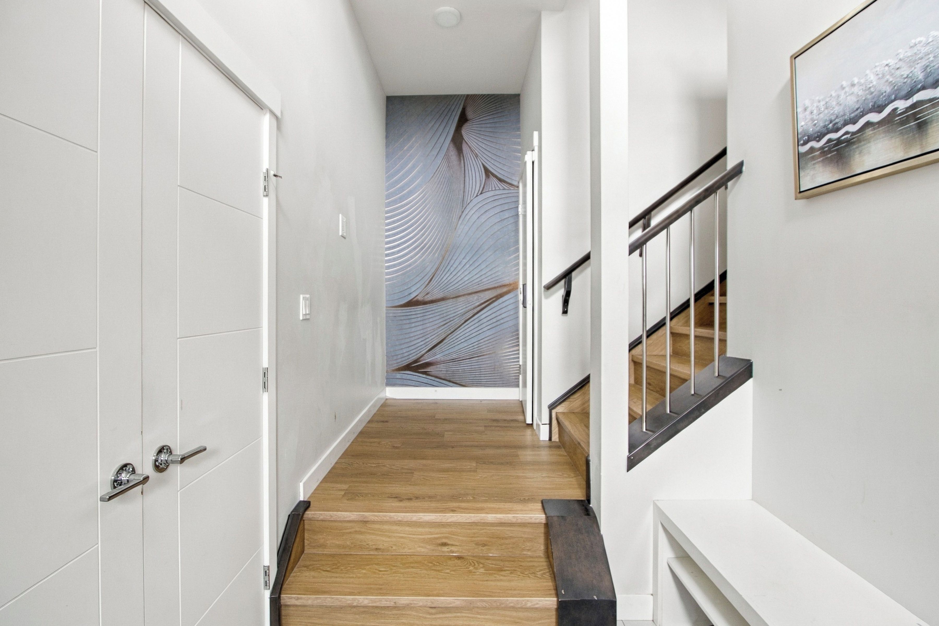 A hallway with a wooden floor, white walls, and a staircase with a metal railing.