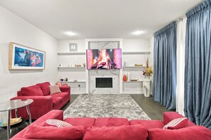 A modern living room with a red sofa set, a flat-screen TV mounted above a fireplace, and a large window with blue curtains. - Spacious Modern Calgary Home | 3 Bedrooms + 2 Bonus Rooms (Calgary)