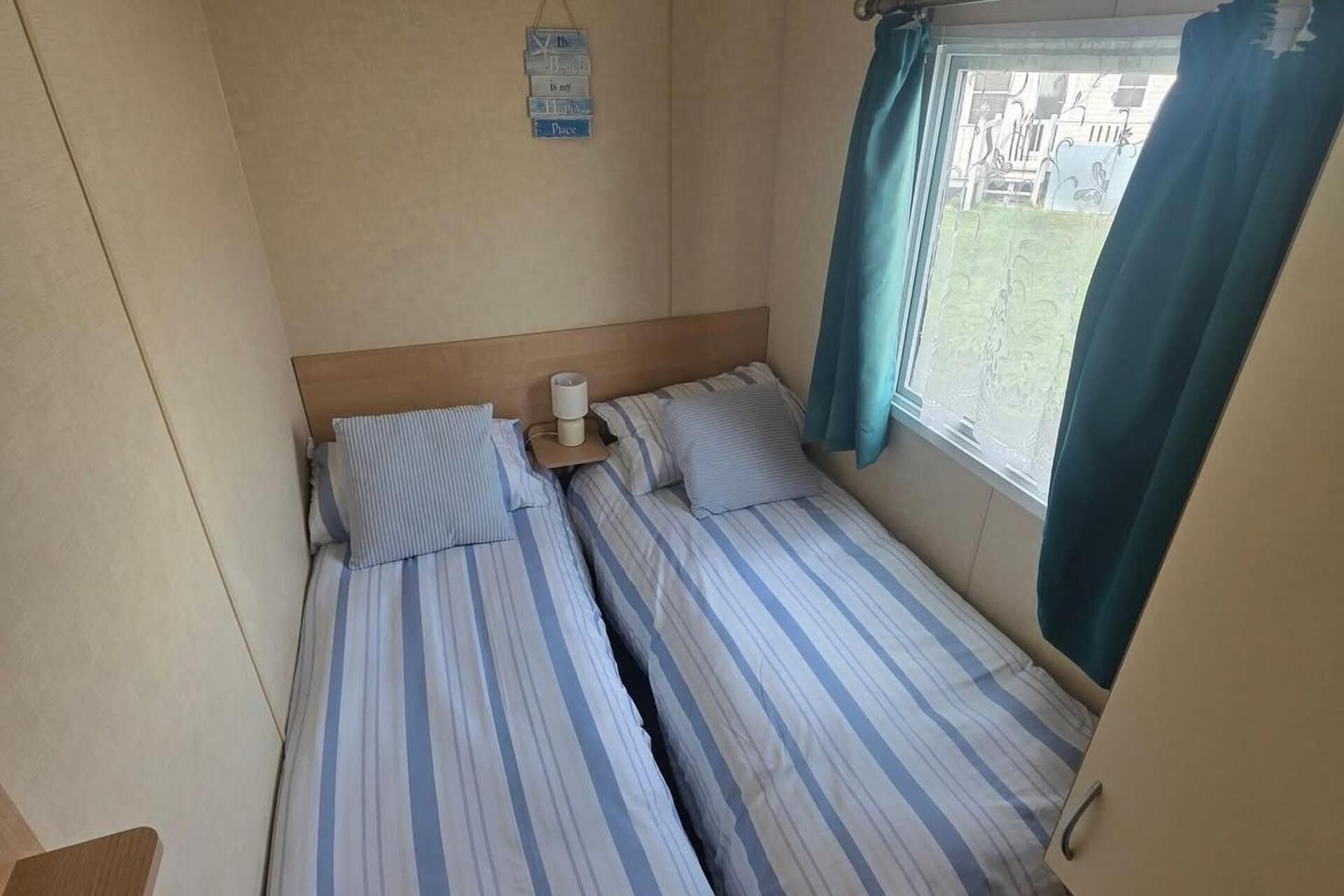 A small, compact room with two beds, a bedside table with a lamp, a window with curtains, and a wall-mounted sign.