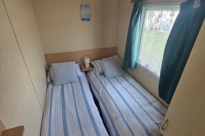 A small, compact room with two beds, a bedside table with a lamp, a window with curtains, and a wall-mounted sign.