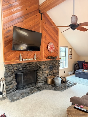 A living room with a stone fireplace, a flat-screen TV mounted on a wooden wall, a ceiling fan, and a cozy seating area.