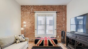 Ein Zimmer mit Backsteinwand, einem Fenster mit Jalousien, einer Couch, einem Fernseher und einer Heizung.