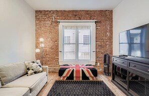 Ein Zimmer mit Backsteinwand, einem Fenster mit Jalousien, einer Couch, einem Fernseher und einer Heizung.
