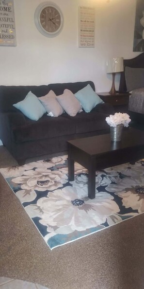 A living room with a dark-colored sofa, a floral-patterned rug, a coffee table, and a wall clock.