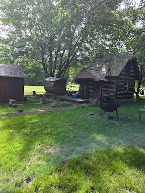 Une cabane rustique en rondins de bois, agrĂ©mentĂ©e dâun tonneau et dâune chaise suspendue, le tout entourĂ© dâune vĂ©gĂ©tation luxuriante.