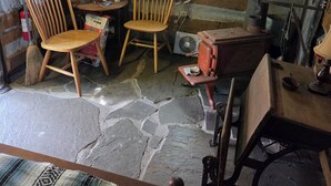 Interior rústico de cabana com paredes de madeira, chão de pedra, duas cadeiras de madeira, uma mesa pequena, uma janela e uma lareira a lenha.