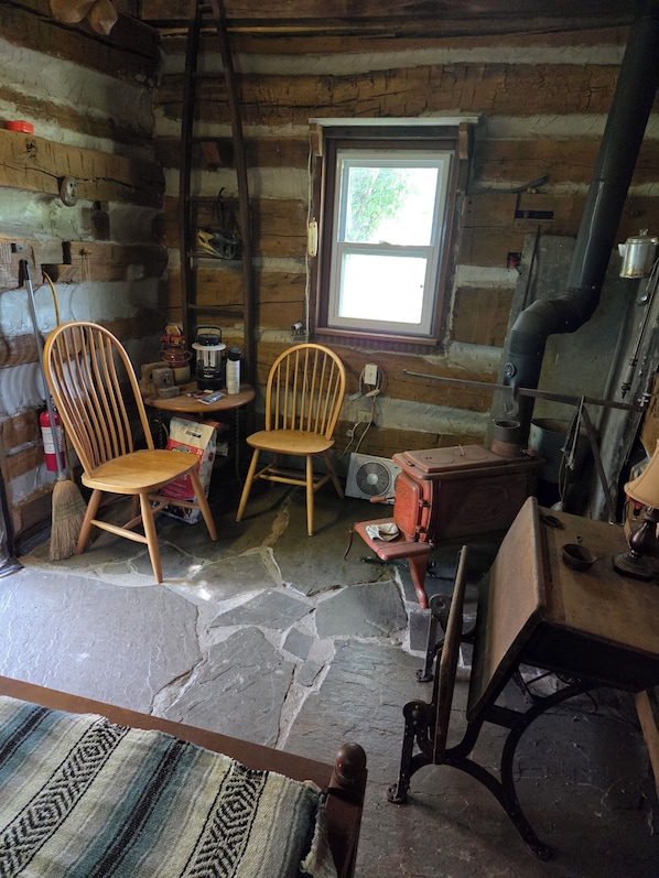 LâintĂ©rieur dâun chalet rustique, avec des murs en bois, un sol en pierre, deux chaises en bois, une petite table, une fenĂȘtre et un poĂȘle Ă bois.
